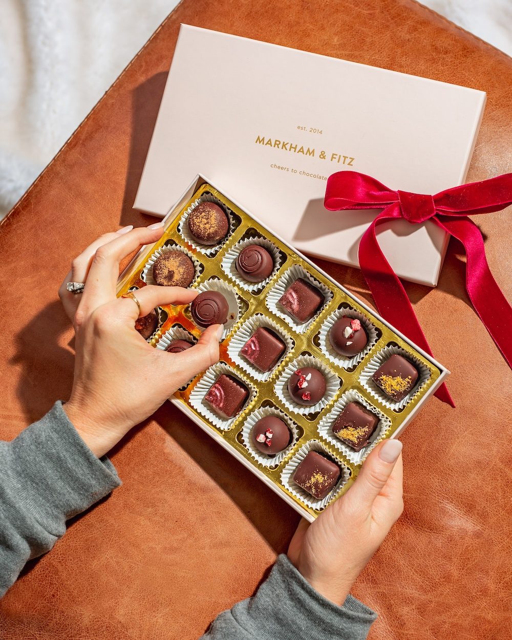 Hands opening a pink Markham & Fitz box of 15 truffles, red velvet ribbon beside it